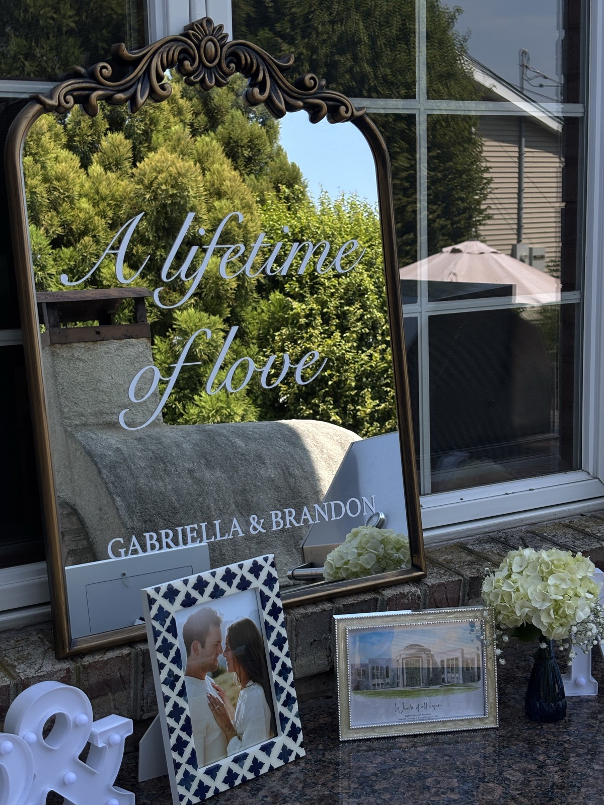 Ornate gold mirror with 'A Lifetime of Love' calligraphy, framed photos, and hydrangea arrangement